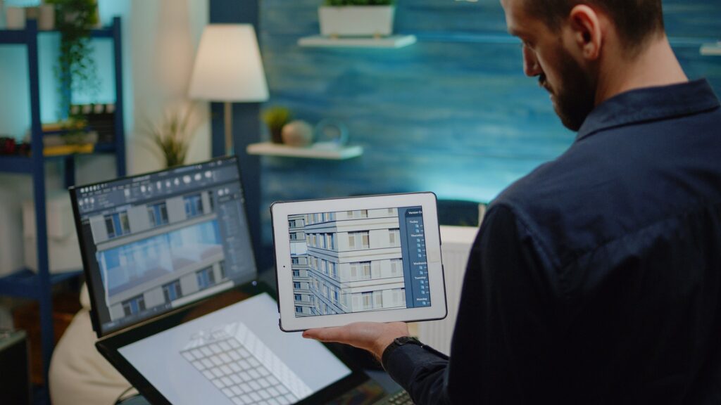 A designer holding a tablet showing a building model beside a laptop