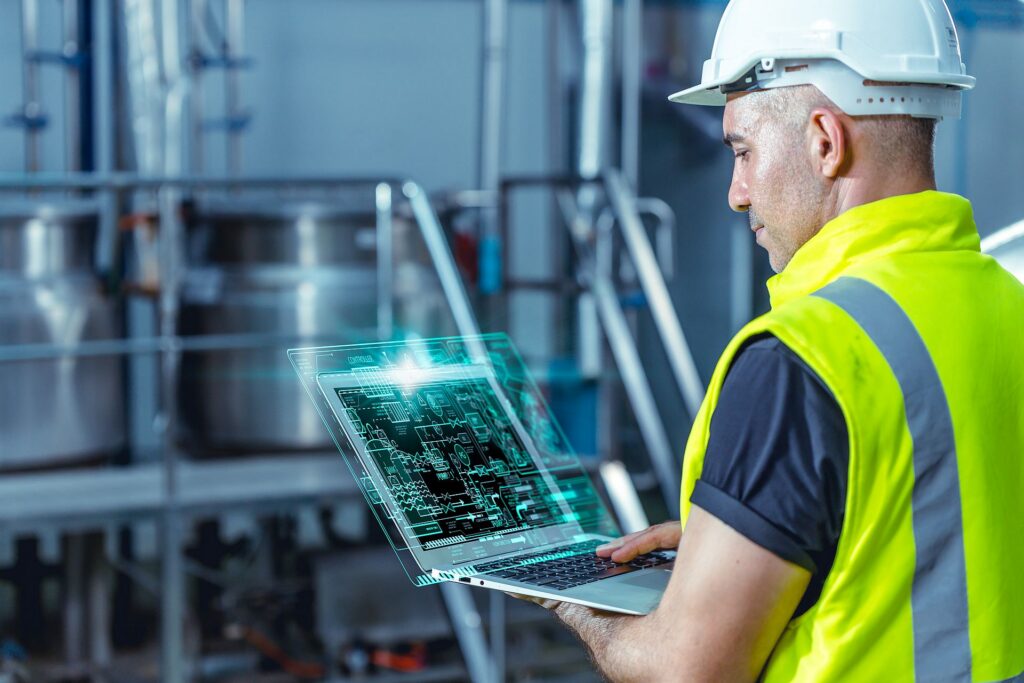 An engineer in a hard hat and high-visibility vest working on a laptop in a plant room